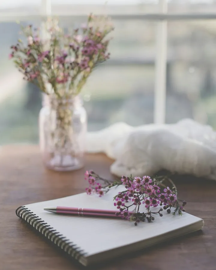 Un carnet à spirale, un stylo et un brin de fleurs mauves sur un bureau en bois, devant une fenêtre
